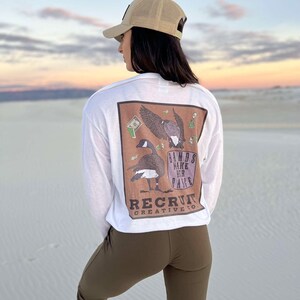 May include: A white long-sleeve t-shirt with a brown graphic of a goose with wings spread and the text "Bands Make Her Dance" and "Recruit Creative Co."