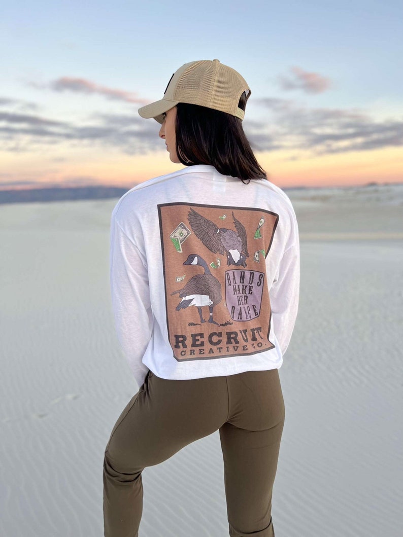 May include: A white long-sleeve t-shirt with a brown graphic of a goose with wings spread and the text "Bands Make Her Dance" and "Recruit Creative Co."