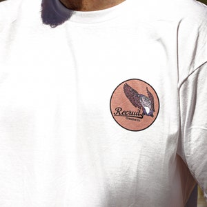 May include: White t-shirt with a brown circle patch featuring a bird in flight and the text "Recruit Creative Co."