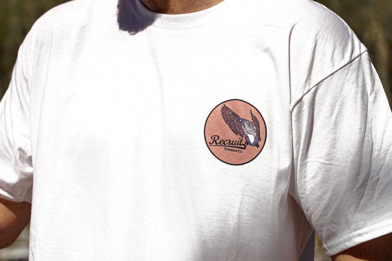 May include: White t-shirt with a brown circle patch featuring a bird in flight and the text "Recruit Creative Co."