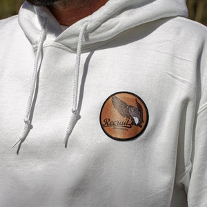 May include: White hooded sweatshirt with a brown circular patch featuring a bird in flight and the text "Recruit Outfitters Co."