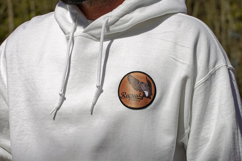 May include: White hooded sweatshirt with a brown circular patch featuring a bird in flight and the text "Recruit Outfitters Co."
