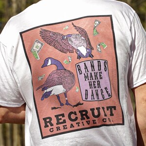 May include: White t-shirt with a pink and orange graphic print of two geese and dollar bills. The text "BANDS MAKE HER DANCE" and "RECRUIT CREATIVE CO." are printed on the graphic.
