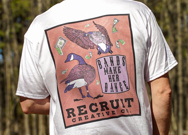 May include: White t-shirt with a pink and orange graphic print of two geese and dollar bills. The text "BANDS MAKE HER DANCE" and "RECRUIT CREATIVE CO." are printed on the graphic.