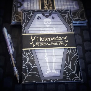 May include: A stack of Halloween-themed notepads with a coffin design, a skeleton illustration, and a spiderweb pattern. The notepads have a black band with the word "Notepads" and "40 sheets Handcrafted". A clear pen is on the left.