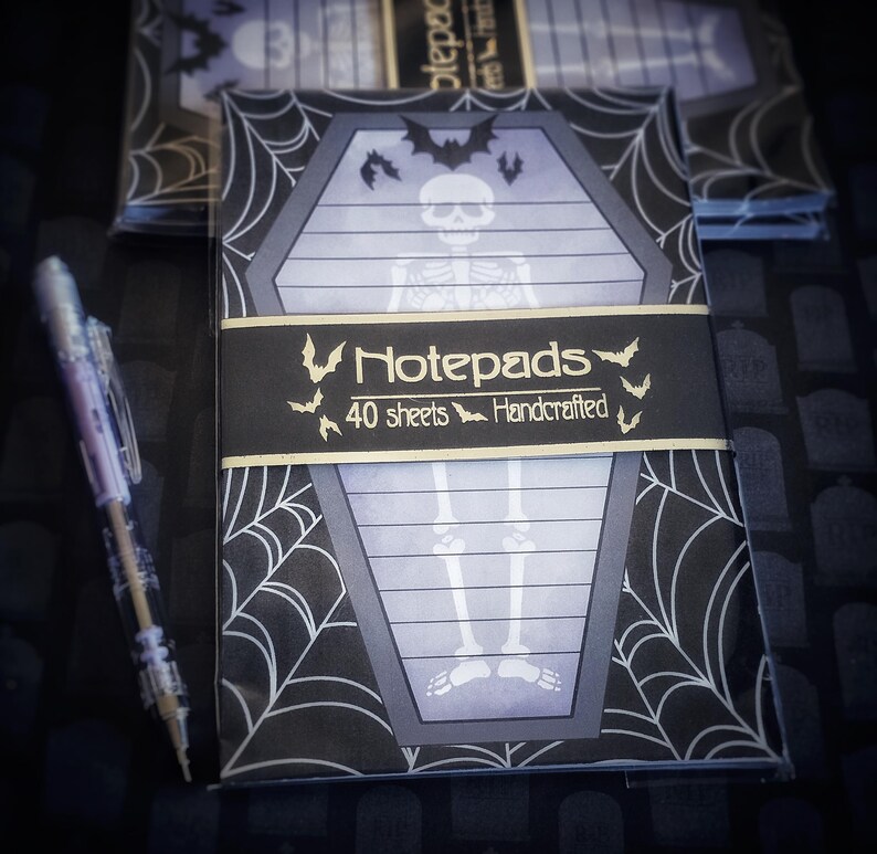 May include: A stack of Halloween-themed notepads with a coffin design, a skeleton illustration, and a spiderweb pattern. The notepads have a black band with the word "Notepads" and "40 sheets Handcrafted". A clear pen is on the left.