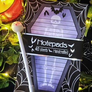 May include: A Halloween-themed notepad with a coffin-shaped design featuring a skeleton illustration. The notepad is wrapped with a black band that reads "Notepads 40 sheets Handcrafted". A white pen with a ghost-shaped topper is in the foreground.