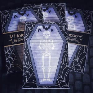 May include: Halloween-themed notepad set. Each notepad features a coffin-shaped design with a skeleton illustration and bat silhouettes. The notepads are surrounded by a spiderweb pattern and include the text "Note 40 sheets" and "pads Handcrafted".