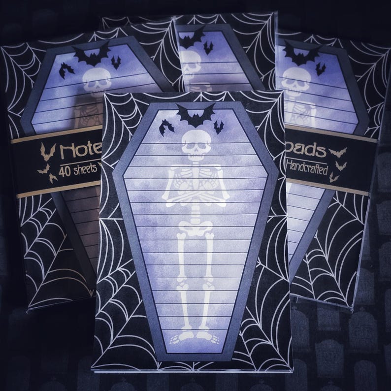 May include: Halloween-themed notepad set. Each notepad features a coffin-shaped design with a skeleton illustration and bat silhouettes. The notepads are surrounded by a spiderweb pattern and include the text "Note 40 sheets" and "pads Handcrafted".