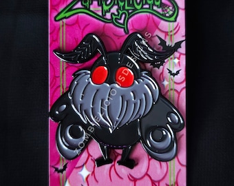 Può includere: Spilla in smalto nero e grigio con una falena di cartone animato con occhi rossi e un corpo peloso. La spilla è su uno sfondo rosa con il testo "Zombielicious Designs" e un indirizzo di sito web.