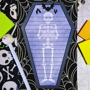 May include: A coffin-shaped notepad with a skeleton illustration and bat silhouettes. The notepad has a blue background with horizontal lines and a dark gray border. The background includes a black and white spiderweb and skull pattern, with colorful sticky notes and a ghost pen.