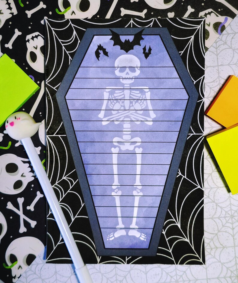 May include: A coffin-shaped notepad with a skeleton illustration and bat silhouettes. The notepad has a blue background with horizontal lines and a dark gray border. The background includes a black and white spiderweb and skull pattern, with colorful sticky notes and a ghost pen.