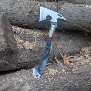 May include: A silver and black axe with a curved handle and a toothed edge on the blade. The axe head has a hammer and a circular cutout. The handle is black with silver accents. The axe is lying on a bed of wood chips and logs.