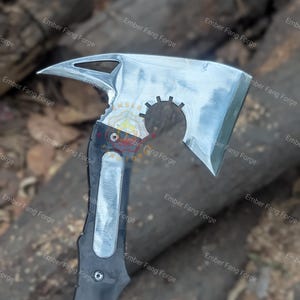 May include: A silver and black axe with a unique design. The axe head has a sharp blade, a pointed end, and a circular cutout. The handle is black with a silver insert. The text "Ember Fang Forge" is visible.
