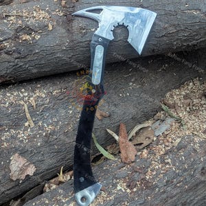 May include: A silver and black tactical axe with a curved blade, a gear-shaped cutout, and a pointed end. The handle is black with silver accents. The axe is resting on a bed of wood shavings and logs.