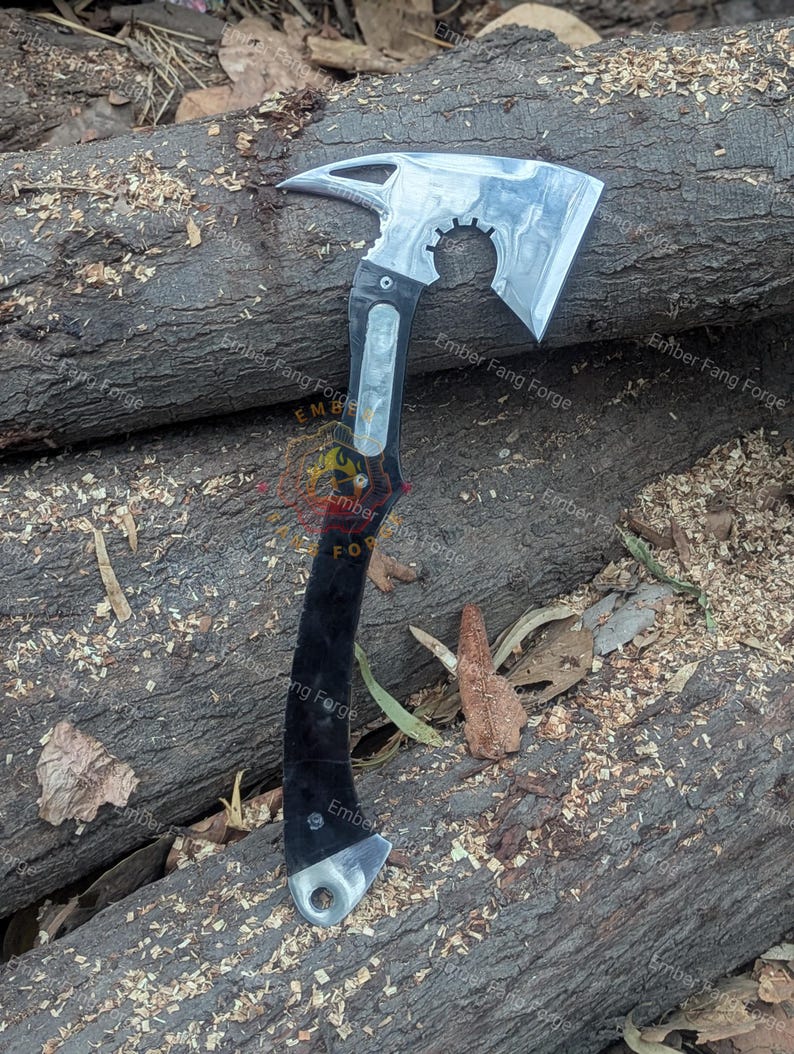 May include: A silver and black tactical axe with a curved blade, a gear-shaped cutout, and a pointed end. The handle is black with silver accents. The axe is resting on a bed of wood shavings and logs.