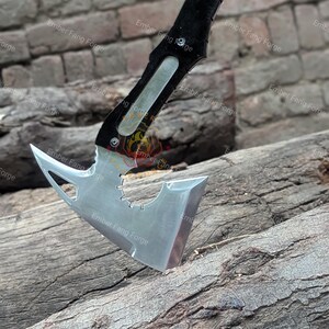 May include: A tactical axe with a polished steel blade and a black handle. The axe head features a unique design with a gear-shaped cutout and a sharp, angled edge. The handle is textured for grip. The axe is resting on a weathered wooden surface.