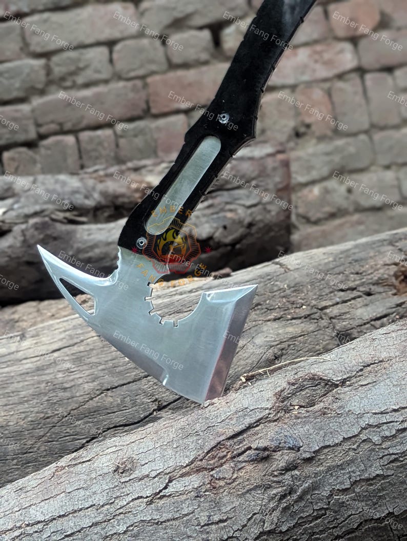 May include: A tactical axe with a polished steel blade and a black handle. The axe head features a unique design with a gear-shaped cutout and a sharp, angled edge. The handle is textured for grip. The axe is resting on a weathered wooden surface.