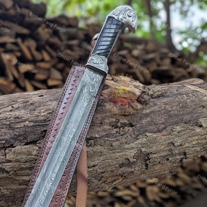 May include: A detailed image of a Damascus steel sword with a decorative silver handle featuring an eagle head design. The sword rests in a brown leather sheath with embossed patterns, showcasing the craftsmanship of the blade and handle.