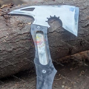 May include: A polished steel axe with a black handle. The axe head features a sharp blade, a pointed spike, and a circular cutout. The handle has a curved design and is riveted to the axe head. The axe is designed for utility and display.