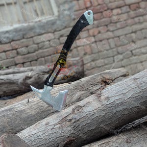 May include: A silver and black hatchet with a curved black handle and a silver head. The hatchet is resting on a pile of wooden logs. The text "Ember Fang Forge" is visible in the background.