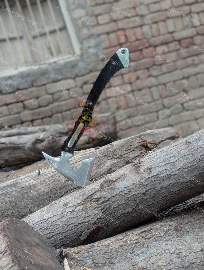 May include: A silver and black hatchet with a curved black handle and a silver head. The hatchet is resting on a pile of wooden logs. The text "Ember Fang Forge" is visible in the background.