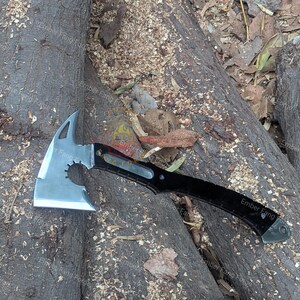 May include: A silver and black tactical axe with a tomahawk-style head. The axe features a serrated edge and a pointed spike on the opposite side of the blade. The handle is black with the text "Ember Fang".