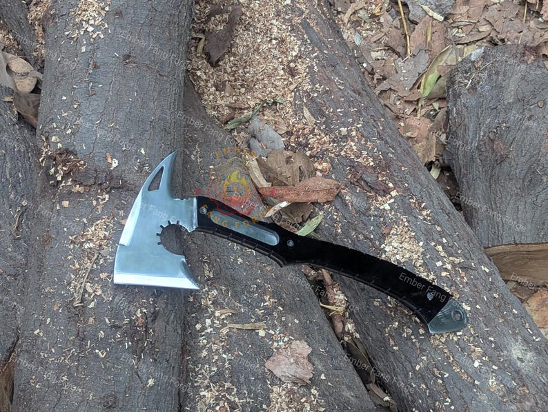 May include: A silver and black tactical axe with a tomahawk-style head. The axe features a serrated edge and a pointed spike on the opposite side of the blade. The handle is black with the text "Ember Fang".