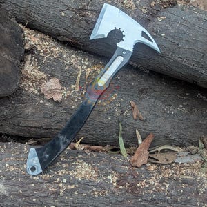 May include: A silver and black axe with a unique design, featuring a toothed wheel and a pointed end. The axe has a black handle and is lying on a bed of wood shavings and logs. The text "Ember Fang Forge" is visible.