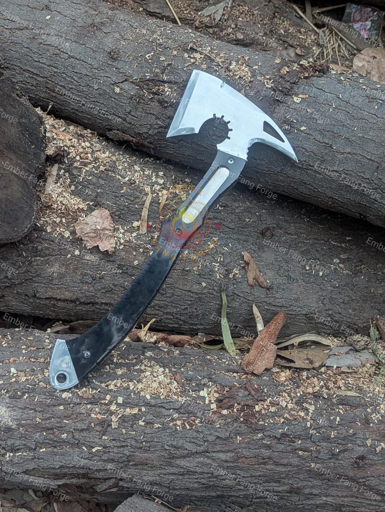 May include: A silver and black axe with a unique design, featuring a toothed wheel and a pointed end. The axe has a black handle and is lying on a bed of wood shavings and logs. The text "Ember Fang Forge" is visible.