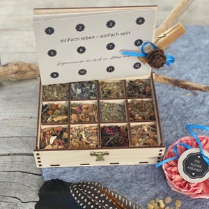 Könnte beinhalten: Eine Holzkiste mit 12 Fächern, gefüllt mit getrockneten Kräutern und Blumen. Eine Karte mit dem Text "einfach leben - einfach sein" ist aufgeklappt. Ein kleiner Tannenzapfen und ein Anhänger mit "DIE KLEINE FREIHEIT" sind mit blauem Band befestigt.
