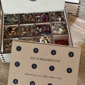 Könnte beinhalten: Eine Holzkiste mit 12 Fächern, gefüllt mit getrockneten Kräutern und Gewürzen. Die Kiste ist mit "Die 12 Rauhnächte" beschriftet und hat die Zahlen 1 bis 12 auf dem Deckel. Die Kiste ist ein Ritual-Set für die 12 Tage Weihnachten.
