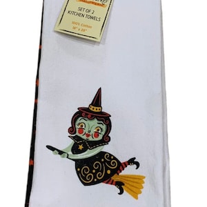 May include: A set of two white 100% cotton kitchen towels measuring 18 inches by 28 inches. The towels feature a graphic of a green-skinned witch with a black hat and dress flying on a broomstick. The tag reads Johanna Parker Halloween.