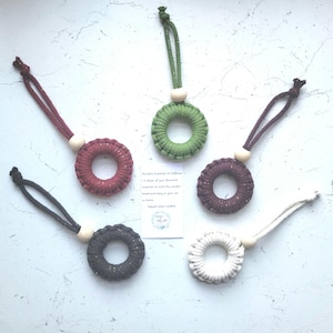 May include: An assortment of macrame essential oil diffusers in shades of burgundy, green, dark grey, and white. Each diffuser features a wooden bead and a loop for hanging. A small card with text is also visible.