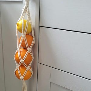 May include: A white macrame hanging fruit basket with a wooden ring at the top. The basket is filled with three pieces of fruit: a lemon and two oranges.