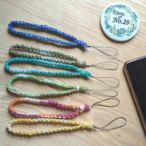 May include: Five colourful macrame wrist straps in blue, green, and yellow hues. Each strap has a loop for attaching to a phone or other device. A round button with the text "Knots No. 29" is also visible.
