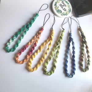 May include: Six handmade phone lanyards in various colours. Each lanyard has a twisted rope design in shades of green, orange, yellow, blue, and brown. The lanyards have a black cord loop for attaching to a phone or other device.