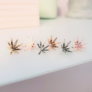 May include: Five gold-toned marijuana leaf earrings in different finishes, including rose gold, silver, and black.