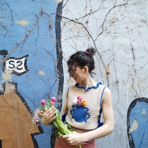 May include: A crocheted sleeveless top with a blue vase and pink, orange, and white flowers. The top is beige with blue trim and a ruffled neckline.