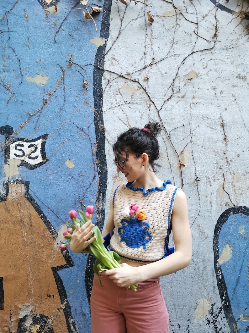 May include: A crocheted sleeveless top with a blue vase and pink, orange, and white flowers. The top is beige with blue trim and a ruffled neckline.