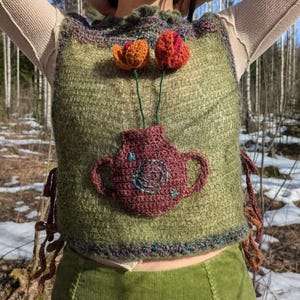May include: A green knitted vest with a brown crocheted vase and three crocheted flowers. The flowers are orange, red, and pink. The vest has a brown crocheted trim around the neckline and the bottom hem. The vest has brown crocheted ties on the sides.