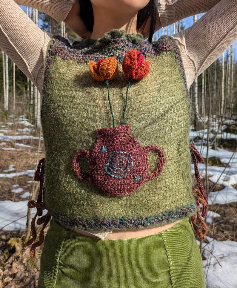 May include: A green knitted vest with a brown crocheted vase and three crocheted flowers. The flowers are orange, red, and pink. The vest has a brown crocheted trim around the neckline and the bottom hem. The vest has brown crocheted ties on the sides.