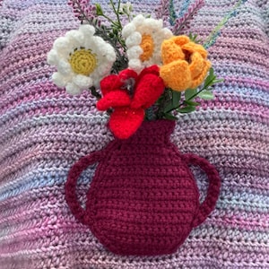 May include: A crocheted sweater with a pink, purple, and blue color gradient. A crocheted vase with a red flower and white flowers is attached to the front of the sweater.