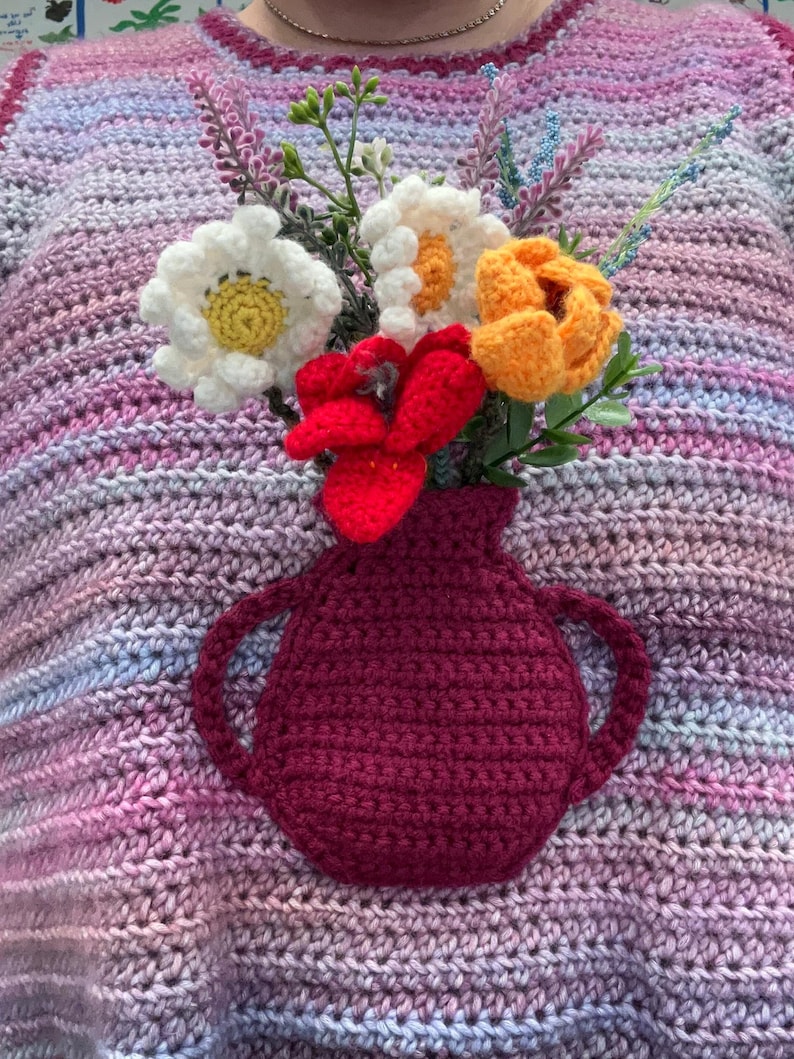 May include: A crocheted sweater with a pink, purple, and blue color gradient. A crocheted vase with a red flower and white flowers is attached to the front of the sweater.