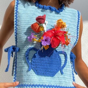 May include: A crocheted sleeveless top with a blue and light blue color scheme. The top features a crocheted vase with a bouquet of flowers in various colors, including red, orange, yellow, purple, and white.