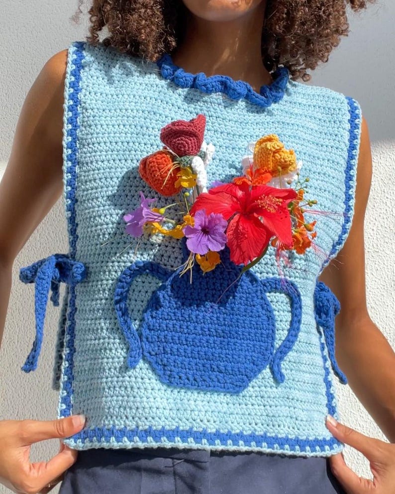 May include: A crocheted sleeveless top with a blue and light blue color scheme. The top features a crocheted vase with a bouquet of flowers in various colors, including red, orange, yellow, purple, and white.