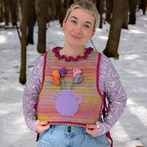 May include: A multi-colored crochet vest with a purple crocheted vase and flowers. The vest has a ruffled neckline and is tied at the sides. The vest is worn over a long-sleeved, purple and white patterned shirt and blue jeans.