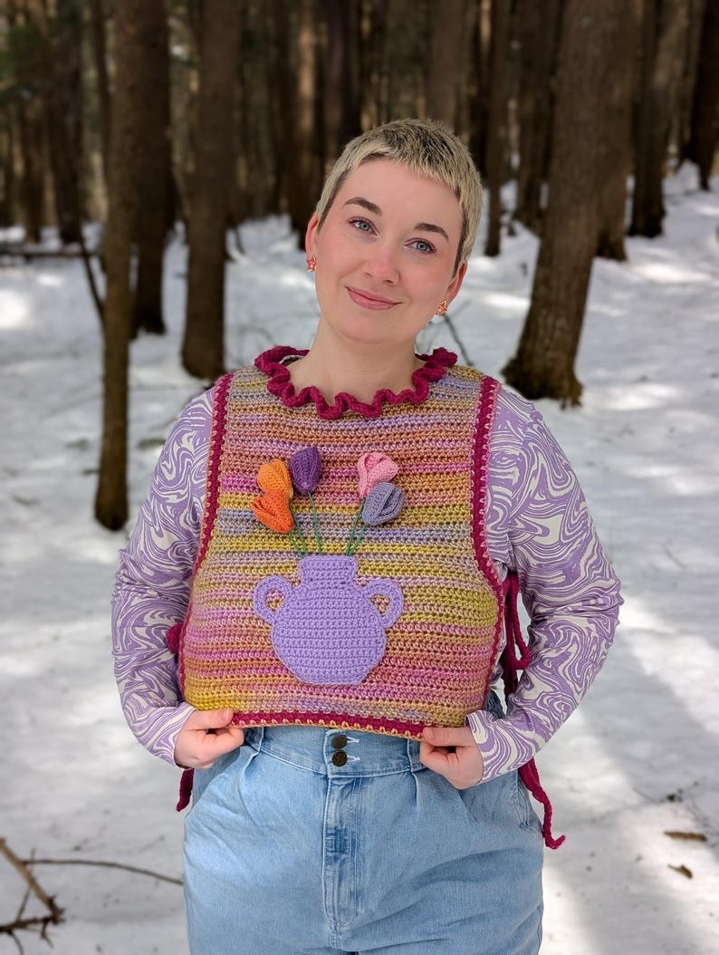 May include: A multi-colored crochet vest with a purple crocheted vase and flowers. The vest has a ruffled neckline and is tied at the sides. The vest is worn over a long-sleeved, purple and white patterned shirt and blue jeans.