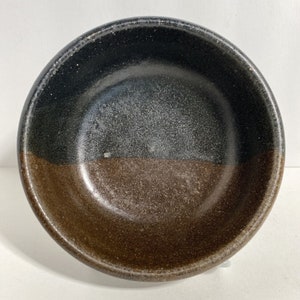 May include: A ceramic bowl with a black and brown speckled glaze. The bowl is divided in half, with the top half being black and the bottom half being brown.