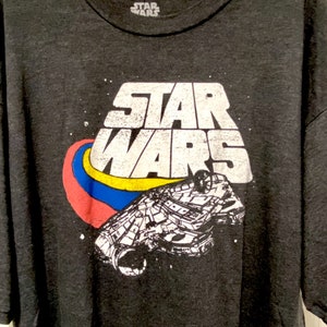 Puede incluir: Una camiseta gris oscuro con un gráfico blanco de las palabras "Star Wars" y una nave espacial. La nave espacial está rodeada por un remolino rojo, azul y amarillo.